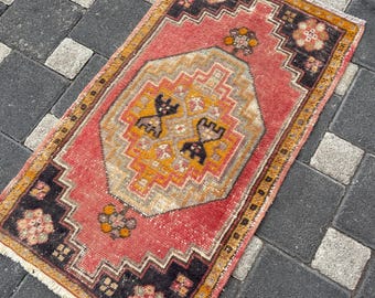 Könnte beinhalten: Ein rechteckiger Teppich mit einem zentralen geometrischen Medaillon in Beige-, Orange- und Schwarztönen auf korallenrotem Grund. Die Bordüre zeigt dunkel anthrazitfarbene Ecken mit Blumenmotiven und Fransen an den oberen und unteren Enden.