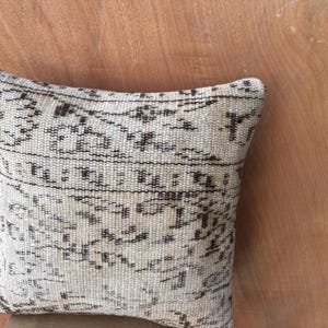 May include: A rectangular cushion with a cream-coloured background and a repeating pattern in dark brown. The cushion has a woven texture and is placed on a wooden surface.
