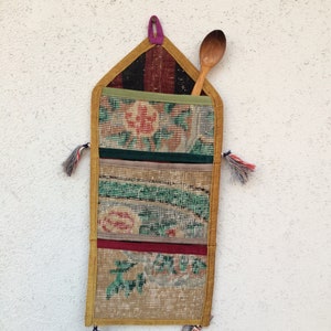 May include: A wall-mounted organizer made from colorful patterned fabric with multiple pockets and tassels. A wooden spoon is hanging in one of the pockets.