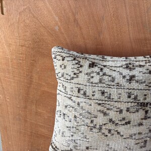 May include: Close-up of a decorative cushion with a cream background and a dark brown, intricate pattern. The cushion's design features geometric shapes and lines, creating a textured appearance. The cushion is placed against a wooden surface.
