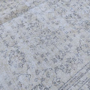 May include: Close-up of a vintage rug with a faded, distressed appearance. The rug features a cream-colored background with intricate floral and geometric patterns in shades of gray and beige. The rug's texture is visible.
