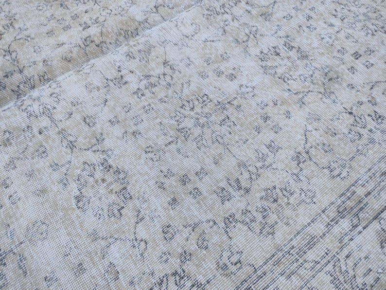 May include: Close-up of a vintage rug with a faded, distressed appearance. The rug features a cream-colored background with intricate floral and geometric patterns in shades of gray and beige. The rug's texture is visible.