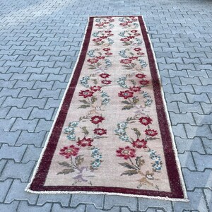 Pu&ograve; includere: Un tappeto rettangolare vintage con un motivo floreale. Il tappeto presenta uno sfondo beige con un motivo ripetuto di fiori rossi e blu. Un bordo rosso scuro incornicia i bordi. Il tappeto &egrave; posizionato su una superficie grigia simile a mattoni.
