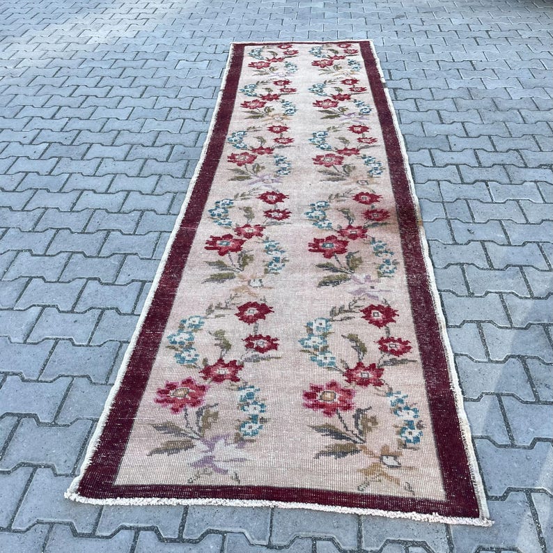 Pu&ograve; includere: Un tappeto rettangolare vintage con un motivo floreale. Il tappeto presenta uno sfondo beige con un motivo ripetuto di fiori rossi e blu. Un bordo rosso scuro incornicia i bordi. Il tappeto &egrave; posizionato su una superficie grigia simile a mattoni.