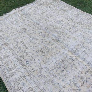 May include: A rectangular, vintage-style rug with a faded floral pattern in cream, beige, and muted blue tones. The rug has a distressed look with a detailed border and fringed edges, suitable for home decor.