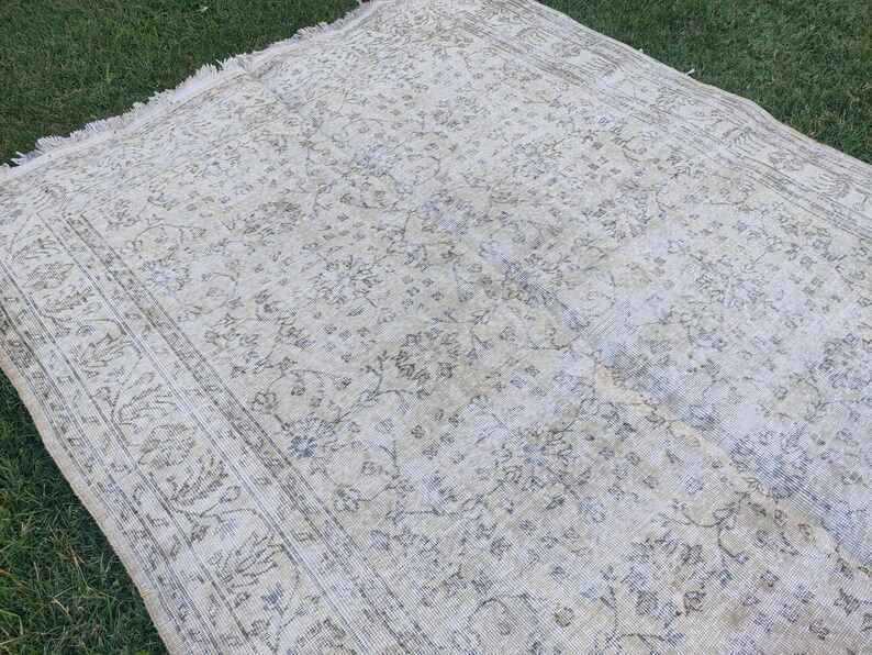 May include: A rectangular, vintage-style rug with a faded floral pattern in cream, beige, and muted blue tones. The rug has a distressed look with a detailed border and fringed edges, suitable for home decor.