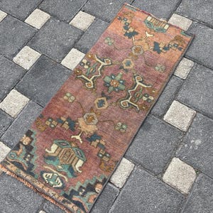 May include: A rectangular woven rug featuring a faded terracotta red base with intricate geometric and floral patterns in shades of teal, dark blue, and tan. The rug has a short fringe at the bottom edge and a dense, low-pile texture throughout.