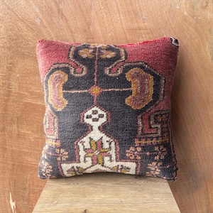 May include: Square cushion with a geometric pattern in shades of red, black, and gold. The cushion has a textured surface and a darker red border. The design features symmetrical shapes and a central figure.