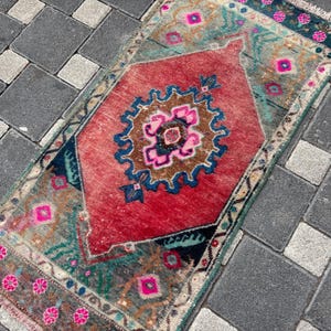 May include: A rectangular woven rug featuring a large central red diamond medallion with a blue, brown, and hot pink floral pattern. The surrounding field is a muted green with additional pink floral motifs and a fringed edge on the short ends.
