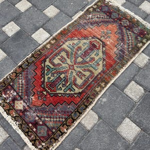 May include: A rectangular woven rug featuring a central geometric medallion in shades of sage green, deep red, and dark blue. The rug has a detailed border with intricate patterns and short white fringe along the top and bottom edges.