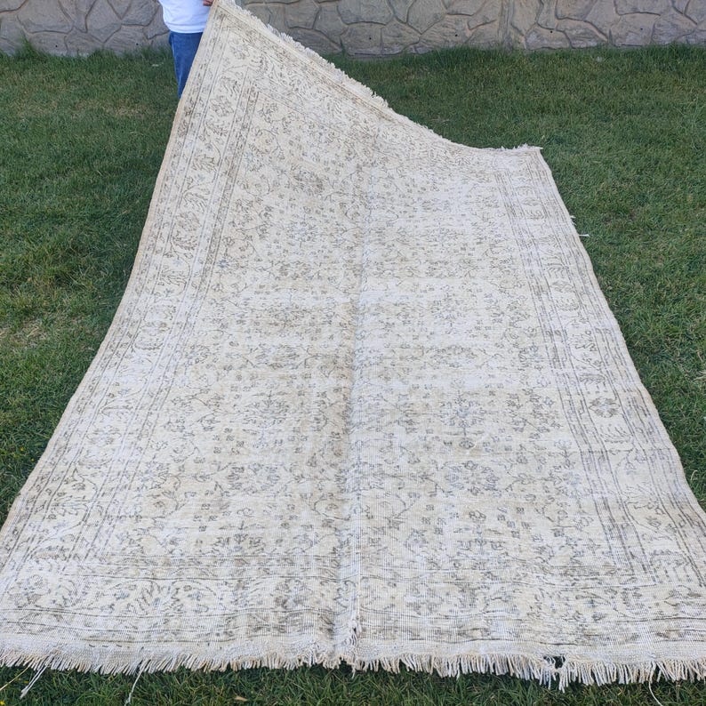 May include: A large, rectangular, vintage-style rug with a cream-colored background and a faded, floral pattern in shades of gray and beige. The rug has a fringed edge and is displayed on green grass.