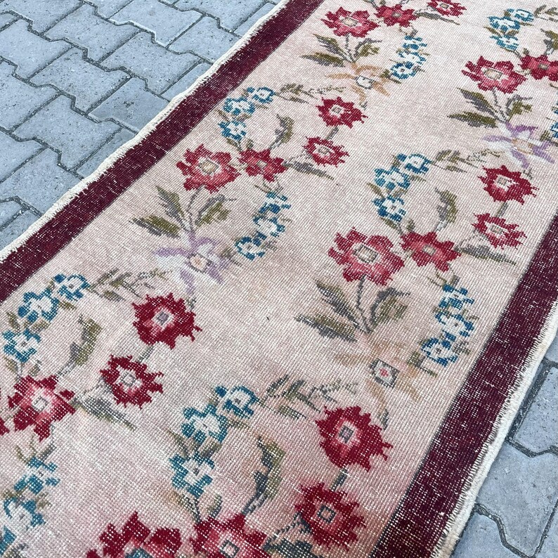 Pu&ograve; includere: Un tappeto vintage con un motivo floreale. Il tappeto presenta uno sfondo rosa chiaro con un motivo ripetuto di fiori rossi e blu. Il bordo &egrave; di colore bordeaux scuro, aggiungendo una cornice a contrasto al design floreale.
