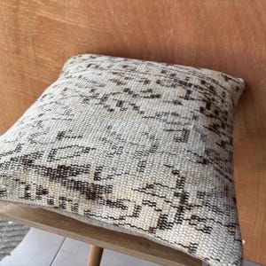 May include: Square beige cushion with a woven, textured design in shades of brown and cream. The cushion is placed on a light wooden chair, showcasing its intricate pattern and neutral colour palette. This decorative cushion adds a touch of rustic charm.