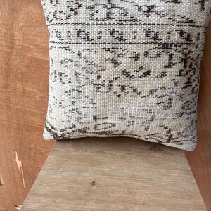 May include: Cream-coloured square cushion with a detailed, dark brown geometric pattern. The cushion is made of a woven fabric and has a textured appearance. The cushion is resting on a wooden surface.