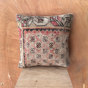 May include: Square cushion with a vintage rug design. The cushion has a beige background with red, black, and yellow geometric patterns. It has a black border, and the overall aesthetic is rustic and textured.