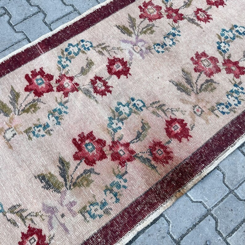 Pu&ograve; includere: Un tappeto rettangolare con sfondo rosa chiaro e motivo floreale. I fiori sono principalmente rossi e blu, con foglie e accenti verdi. Il tappeto ha un bordo rosso scuro.