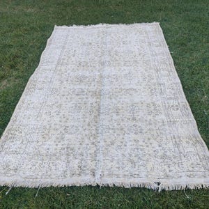 May include: A rectangular, off-white rug with a faded floral pattern and a fringed edge. The rug is laid out on green grass, showcasing its vintage aesthetic and intricate design. The rug's dimensions are not visible.