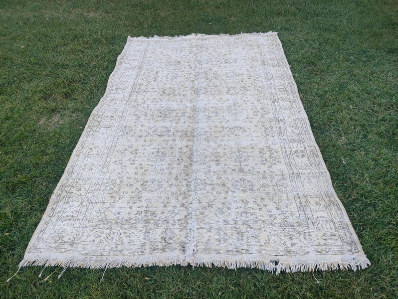 May include: A rectangular, off-white rug with a faded floral pattern and a fringed edge. The rug is laid out on green grass, showcasing its vintage aesthetic and intricate design. The rug's dimensions are not visible.