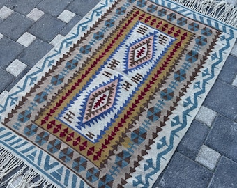 Puede incluir: Una alfombra tejida rectangular con una base de color crema y un patrón geométrico en tonos azul, amarillo mostaza y rojo oscuro. El diseño incluye motivos de diamantes centrales y un borde con flecos en ambos extremos de la pieza.