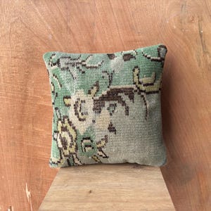 May include: Square cushion with a faded, vintage-style pattern. The cushion features a muted green background with floral and geometric designs in shades of brown and yellow. The cushion is placed on a wooden surface.