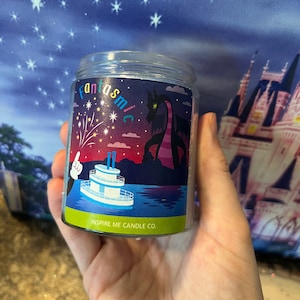 May include: A glass candle jar with a colorful label featuring a steamboat, fireworks, and a dragon. The label reads "Fantasmic" and "Inspire Me Candle Co."