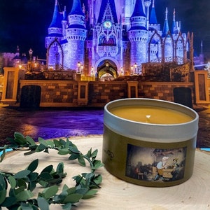 May include: A gold candle with a brown label featuring a cartoon illustration of a person in a blue shirt and a white hat. The candle is sitting on a wooden surface with a green garland in the foreground. The background is a blurry image of a blue and purple castle.