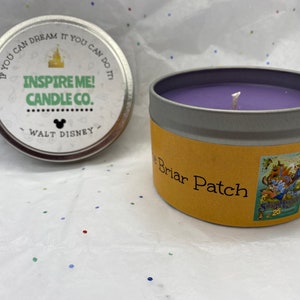 Puede incluir: Una vela morada en una lata plateada con una etiqueta amarilla que dice "The Briar Patch". La tapa de la lata de la vela tiene el texto "If you can dream it you can do it" y "Inspire Me! Candle Co."