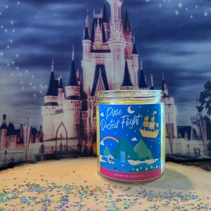 May include: A glass candle jar with a yellow lid and a blue and white label that says "Pixie Dusted Flight" with a cartoon image of a ship flying over a green island. The candle jar is sitting on a wooden surface with silver glitter stars.