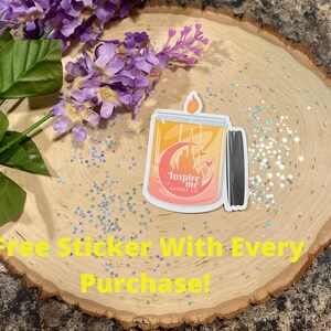 May include: A sticker of a candle with a pink, orange, and yellow ombre design. The candle has a black lid and a white label that says "Inspire Me Candle Co.". The sticker is on a wooden surface with silver glitter.