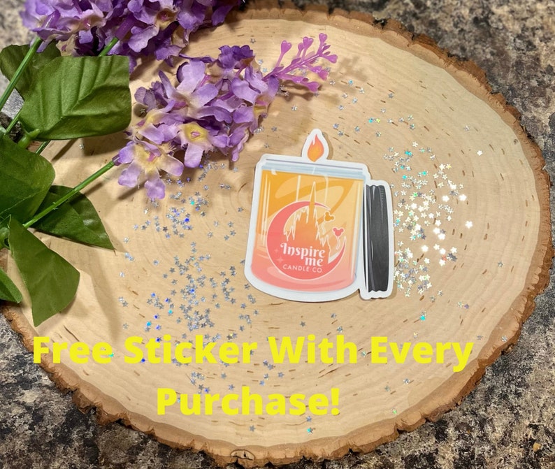 May include: A sticker of a candle with a pink, orange, and yellow ombre design. The candle has a black lid and a white label that says "Inspire Me Candle Co.". The sticker is on a wooden surface with silver glitter.