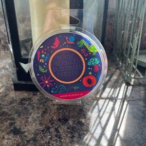 May include: A round, clear candle jar with a colorful label featuring a whimsical illustration of a castle, a snail, a dragon, and mushrooms. The label reads "Main Street Electrical Parade" and "Inspire Me Candle Co."