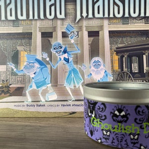May include: A purple and white candle tin with a spooky design featuring the text "Ghoulish Delight". The tin is sitting in front of a book with a picture of a haunted mansion and ghosts.