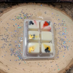 May include: Six wax melts in a clear plastic tray. The melts are white with different colored designs and Mickey Mouse silhouettes. The designs include red, blue, gold, and black.