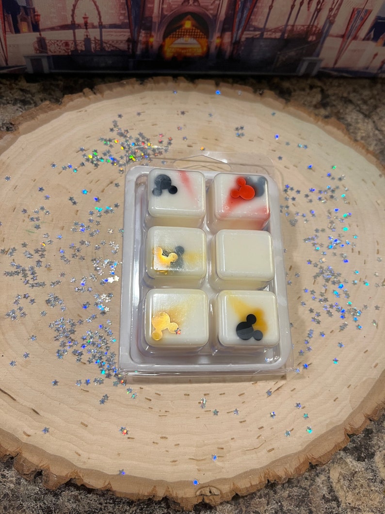 May include: Six wax melts in a clear plastic tray. The melts are white with different colored designs and Mickey Mouse silhouettes. The designs include red, blue, gold, and black.