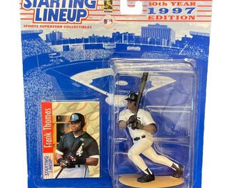 Pode incluir: Uma figura de basebol Starting Lineup de 1997 de Frank Thomas numa caixa de plástico transparente. A embalagem inclui um cartão de basebol e o texto "Starting Lineup" e "10th Year 1997 Edition". A figura está vestida com um uniforme branco e preto.