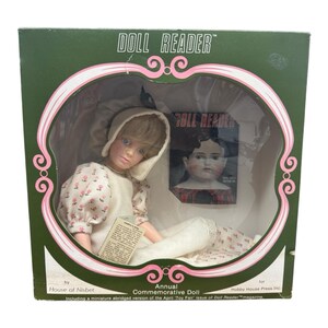 Pode incluir: Uma boneca vintage em uma caixa verde com o texto "DOLL READER". A boneca está usando um capuz branco e um vestido floral. Uma revista em miniatura está incluída. A caixa diz "Annual Commemorative Doll".
