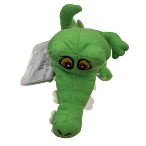 May include: A green plush alligator toy with large, expressive eyes and a detailed snout. The toy has a tag attached and is made of soft, textured fabric. The alligator's body is a vibrant green, with lighter green accents.