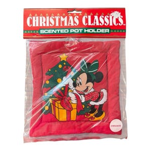 May include: A square red scented pot holder in clear plastic packaging labelled CHRISTMAS CLASSICS. The pot holder features a graphic of Minnie Mouse wearing a green bow, standing next to a yellow gift box and a green Christmas tree.