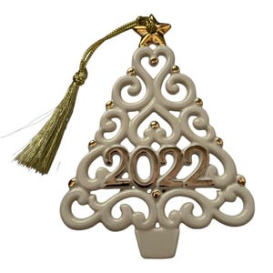 May include: A cream-colored ceramic Christmas tree ornament featuring an intricate openwork scroll design. The center displays the year 2022 in metallic gold. A gold star sits at the top, and a gold tassel hangs from a matching cord.