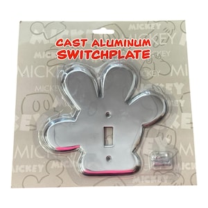 May include: A cast aluminum switchplate in the shape of Mickey Mouse's hand. The switchplate is silver with a pink accent and includes the text "CAST ALUMINUM SWITCHPLATE". The switchplate is packaged on a gray background.