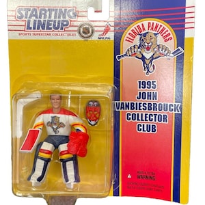 May include: A plastic action figure of a hockey goalie wearing a white, red, and blue uniform with a Florida Panthers logo. The figure is holding a hockey stick and is standing on a yellow background. The packaging includes the text "1995 John Vanbiesbrouck Collector Club".