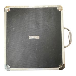 Puede incluir: Una maleta cuadrada negra con marco y asa de metal plateado. La maleta tiene una superficie texturizada y la palabra "MULTIZ" está impresa en una pequeña placa plateada.