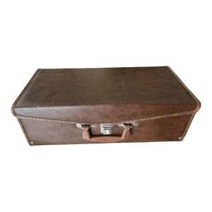 May include: A rectangular brown suitcase with a smooth, solid-colored exterior. The case features a central handle and a silver-toned metal latch mechanism positioned directly above the handle for secure closure of the lid.