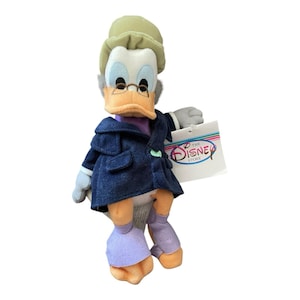 May include: A plush figure of a duck wearing a navy blue coat, a green hat, and glasses. The duck has an orange beak and feet, with light blue eyes and gray hair. The figure is holding a tag that says "The Disney Store".