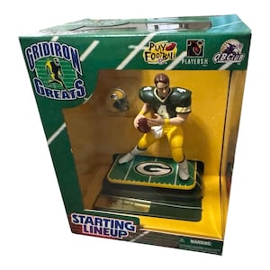 Op de afbeelding: Een Starting Lineup-figuur van een American footballspeler in een groen en geel uniform, met een football. De figuur staat op een basis met een American footballveld-ontwerp. De doos leest "Gridiron Greats" en "Starting Lineup".