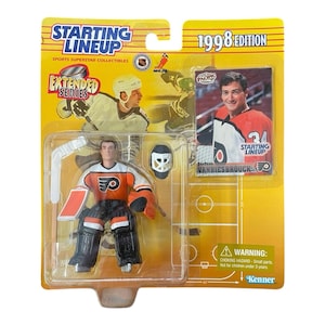 May include: A 1998 edition Starting Lineup action figure of a Philadelphia Flyers hockey goalie, packaged on a yellow and clear plastic card. The figure is wearing black and orange gear. Includes a hockey stick, mask, and a trading card of John Vanbiesbrouck.