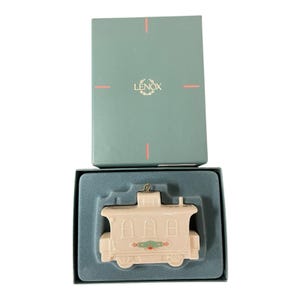 May include: A cream-coloured Lenox Christmas train carriage ornament, presented in its original teal box. The ornament features red and green detailing. The box has the Lenox logo.