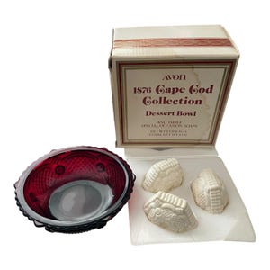May include: A ruby red glass dessert bowl with a textured diamond pattern and a white cardboard box labelled Avon 1876 Cape Cod Collection Dessert Bowl and Three Special Occasion Soaps, including three white soaps with a moulded decorative design.