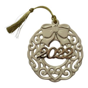 May include: A cream-colored ceramic ornament featuring an intricate openwork scroll design, a sculpted bow at the top, and the year 2022 in metallic gold lettering. A gold-colored cord with a matching tassel is attached to the top loop.