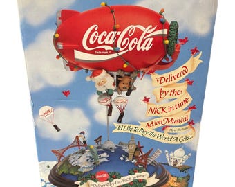 May include: A Coca-Cola themed musical Christmas decoration featuring a red blimp with Santa Claus and a reindeer hanging from it. The blimp is flying over a globe with landmarks from around the world. The box reads "Delivered by the NICK in time" and "I'd Like To Buy The World A Coke."